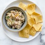 A bowl of salad with canned salmon and chopped vegetables sits on a plate next to several square, yellow crackers on a white marble surface.