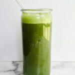 A tall glass of Blender Green Juice topped with foam, a clear straw inserted, sits on a marble surface against a white background.