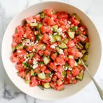 A white bowl filled with a refreshing Watermelon Salad, featuring diced watermelon, cucumber, fresh herbs, and crumbled feta cheese, with a spoon resting inside. The bowl sits atop a white marble surface.