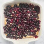 A square white baking dish filled with Blueberry Dump-It Cobbler, its golden surface crowned with a layer of whole, juicy blueberries on a marble countertop.