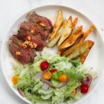 A white plate with sliced steak topped with cherry pepper butter and seasoning, golden potato wedges, and a fresh salad of lettuce, cherry tomatoes, and red onion on a white marble background.