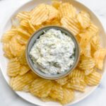 A plate of ridged potato chips arranged around a small bowl of creamy Zucchini Tzatziki Dip with green herbs, set on a white marble surface.