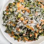 A Tuscan Kale Salad featuring shredded cheese, walnut pieces, and sliced apples is served on a white plate against a marble background.