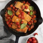 A black skillet filled with Citrus Salmon fillets, cherry tomatoes, and basil leaves, with more cherry tomatoes and fresh basil nearby on a light countertop. A striped towel and plates with forks are also visible.