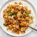 A white plate with caramelized curried cauliflower florets topped with chopped herbs, served over a layer of creamy white sauce, with a spoon resting on the side.