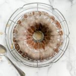 A One-Bowl Banana Bundt Cake sits on a round wire cooling rack over a white marble surface, with a spoon and some dripped glaze nearby.