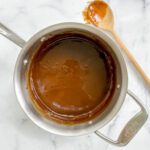 A saucepan filled with smooth salted caramel sauce sits on a white marble surface, with a wooden spoon coated in caramel resting nearby.