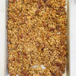 A baking sheet filled with homemade Pumpkin Spice Granola, featuring clusters of oats and pecans, spread evenly and baked to a golden brown. The sheet rests on a white marble surface.