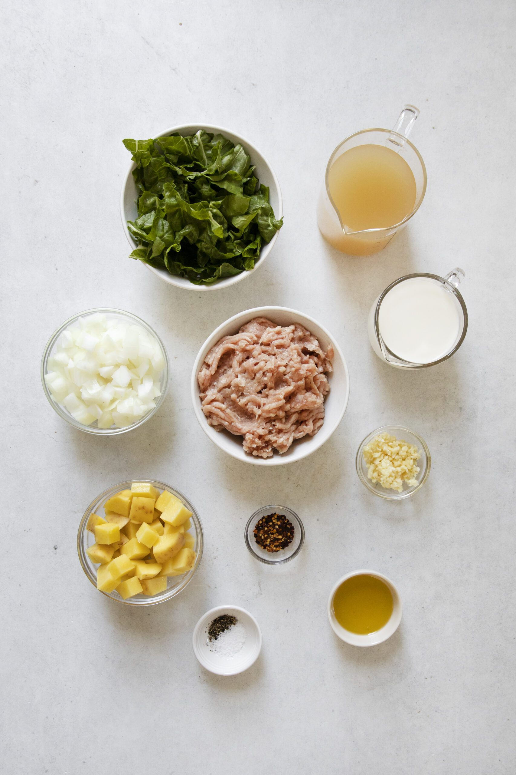 A flat lay of Chicken Potato Soup ingredients on a white surface, including chopped spinach, chicken broth, milk, diced onions, ground meat, minced garlic, diced potatoes, olive oil, black pepper, and red pepper flakes.