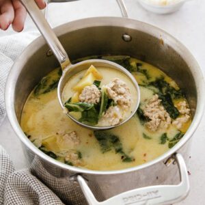 A hand holds a ladle scooping creamy Chicken Potato Soup with sausage, spinach, and pasta from a large metal pot. The soup sits on a light countertop with a checkered towel and small bowl in the background.