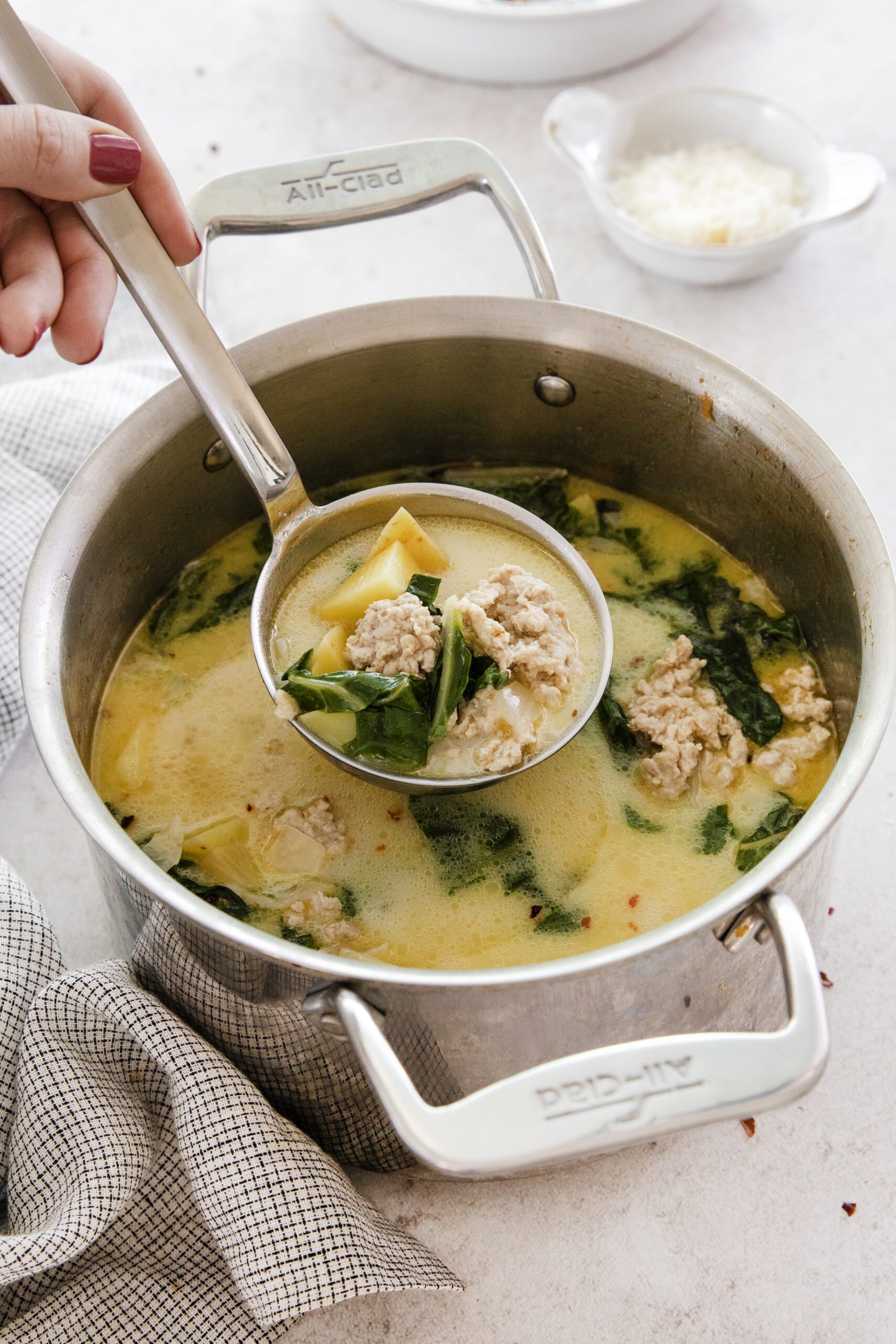 A hand holds a ladle scooping creamy Chicken Potato Soup with sausage, spinach, and pasta from a large metal pot. The soup sits on a light countertop with a checkered towel and small bowl in the background.