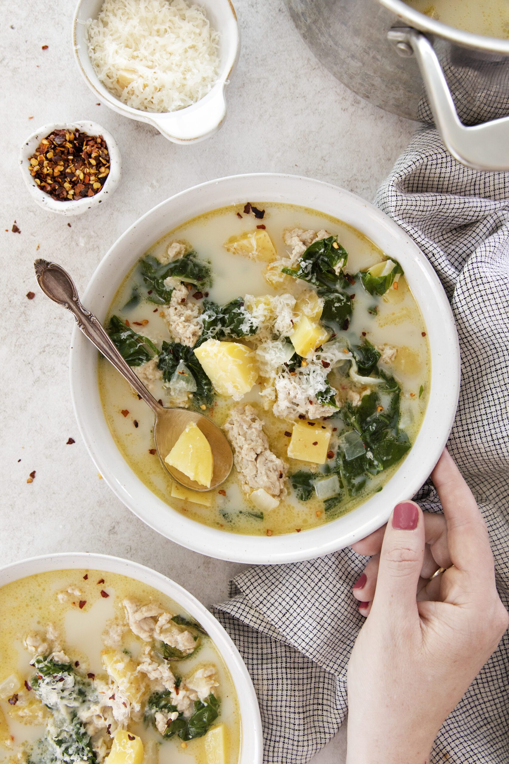 A hand holds a bowl of creamy Chicken Potato Soup with sausage, kale, and grated cheese. Nearby are bowls of grated cheese and red pepper flakes, and a pot of soup on a light surface with a checkered napkin.