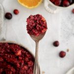 A hand holds up a spoonful of vibrant cranberry relish over a bowl, with fresh cranberries and a halved orange in the background on a light surface.