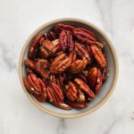 A ceramic bowl filled with Miso-Maple Glazed Pecans sits on a white marble surface.