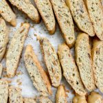 Golden brown Gluten-Free Biscotti with visible almond slices are arranged on a sheet of parchment paper. The cookies have a crisp texture, and some crumbs are scattered around them.