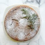 A round Grapefruit Olive Oil Cake dusted with powdered sugar, garnished with sprigs of rosemary, sits on a white plate atop a marble surface.