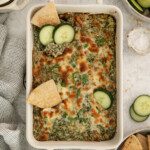 A baked Spinach Dip in a rectangular dish, topped with melted cheese, sliced cucumbers, and pita chips. Nearby are bowls of pita chips, cucumber slices, and salt on a light-colored surface.