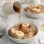 A hand pours syrup from a small white pitcher onto a bowl of slow cooker oatmeal topped with banana slices and walnuts; another bowl of oatmeal is in the background.