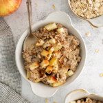A bowl of slow cooker oatmeal topped with chopped apples and sprinkled with cinnamon, with a spoon in the bowl. Nearby are a fresh apple, a bowl of oats, a small dish of cinnamon, and a checkered cloth.
