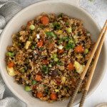 A bowl of vegetable fried rice features chopped carrots, peas, mushrooms, green onions, sesame seeds, and scrambled eggs, served with chopsticks on the side.