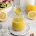 A hand holding a spoon drizzles yellow Lemon Vinaigrette into a glass jar. Sliced lemons, a salad bowl, and a honey dipper rest on the light surface nearby.