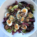 A white bowl filled with leafy greens, sliced boiled eggs, cherry tomatoes, walnuts, and herbs creates a vibrant Fall Salad, beautifully arranged on a marble surface.
