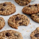 A group of Oatmeal Chocolate Chip Cookies are arranged on a light surface. One cookie in the center has a bite taken out, with scattered oats and a glass of milk in the background.