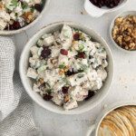 A bowl of creamy Cranberry Turkey Salad with walnuts and herbs sits beside small dishes of dried cranberries, walnuts, and round crackers on a light surface with a checkered cloth nearby.