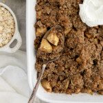 A baking dish filled with flourless apple crisp, topped with oat crumble and a dollop of whipped cream. A spoon holds a portion, showing spiced apple chunks. Nearby are a bowl of oats, a green apple, and a white cloth.