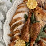 Herb butter roast turkey sliced and arranged on a white platter with two whole drumsticks, fresh sage, and halved lemons for garnish. A striped towel is partially visible at the edge.