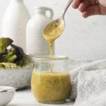 A hand holds a spoonful of creamy yellow Olive Garden Italian Dressing above a glass jar, with some dressing dripping back in. Nearby are a bowl of lettuce, a checkered cloth, and two ceramic bottles in the background.