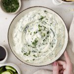 A bowl of creamy swiss chard garlic yogurt dip garnished with chopped chives, surrounded by cucumber slices, chips, Worcestershire sauce, and extra chives. A hand rests on the edge of the bowl.