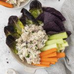 A bowl with tuna salad, purple lettuce, celery sticks, carrot sticks, and blue corn tortilla chips. Nearby are a smaller bowl of Deli-Style Chicken Salad, a bowl of chopped herbs, and more veggie sticks on a light surface with a checkered cloth.