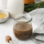A glass jar filled with Honey and Dill Balsamic Dressing sits on a light surface, surrounded by a honey dipper, fresh dill, a checkered cloth, a ceramic jug, and a bottle of balsamic vinegar.