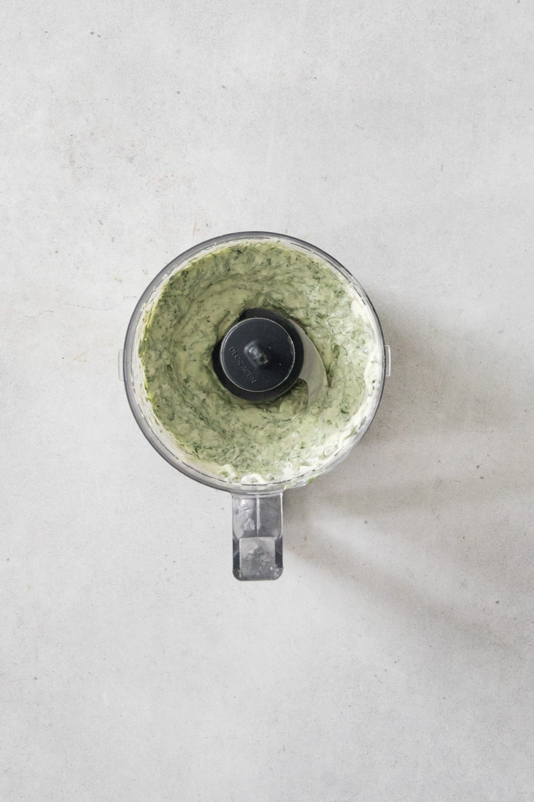 A food processor containing a creamy, green herb mixture is placed on a light gray countertop, viewed from above.