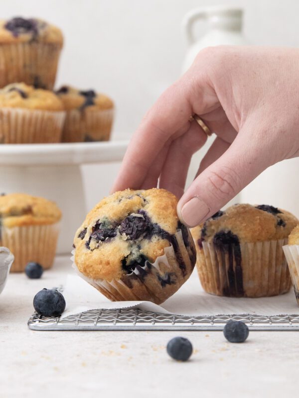 A hand picks up one of the freshly baked blueberry muffins from a cooling rack. Other blueberry muffins and loose blueberries are scattered nearby, with even more stacked elegantly on a cake stand in the background.