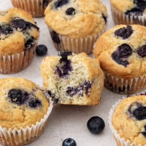 A group of blueberry muffins sits on a light surface, with whole blueberries scattered around. One blueberry muffin in the center is cut open, revealing its soft, moist interior studded with juicy blueberries.