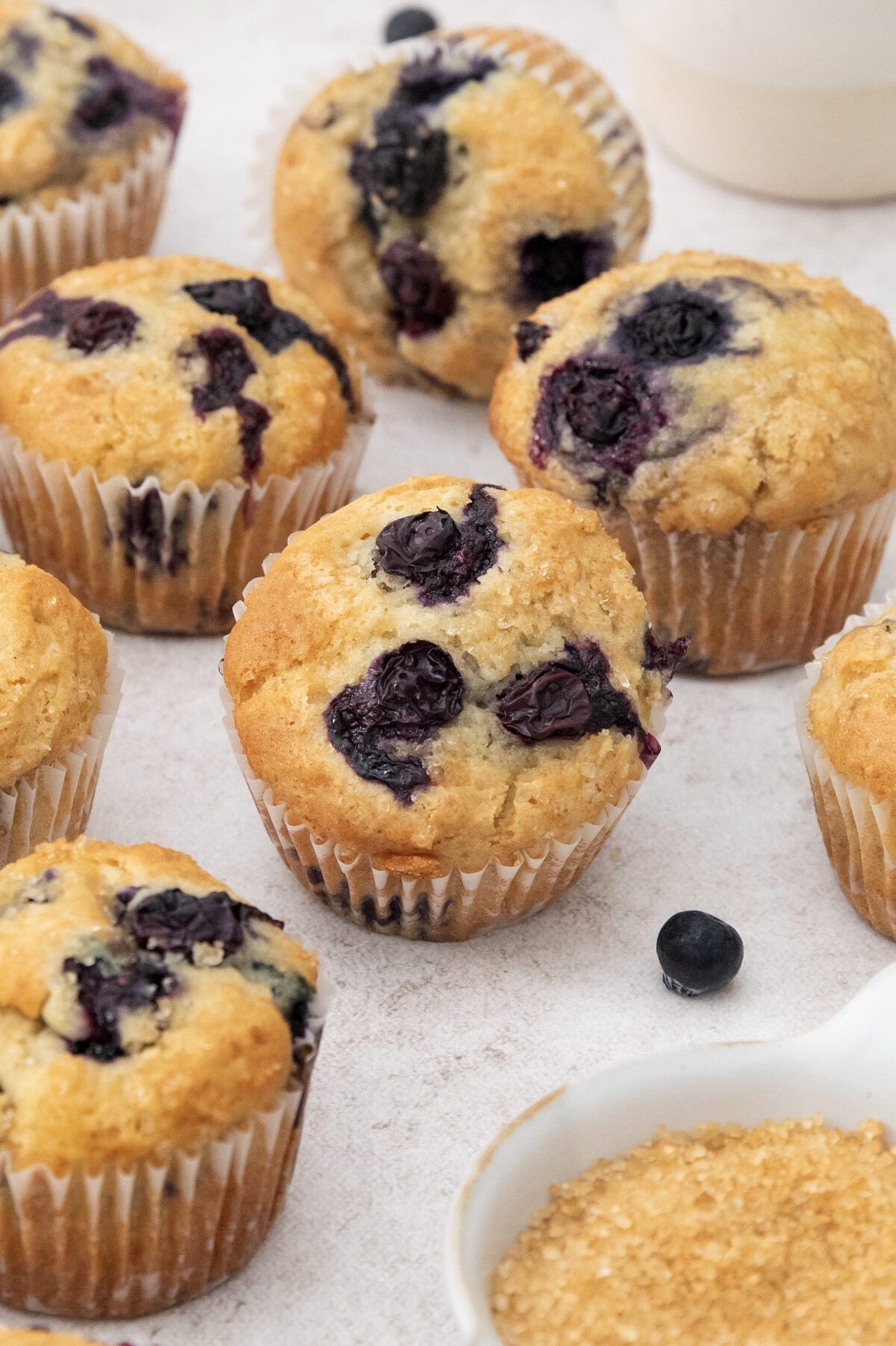 Several golden brown blueberry muffins are arranged on a light surface, with juicy blueberries baked inside. A small bowl of coarse sugar and a few loose blueberries surround the freshly baked blueberry muffins.