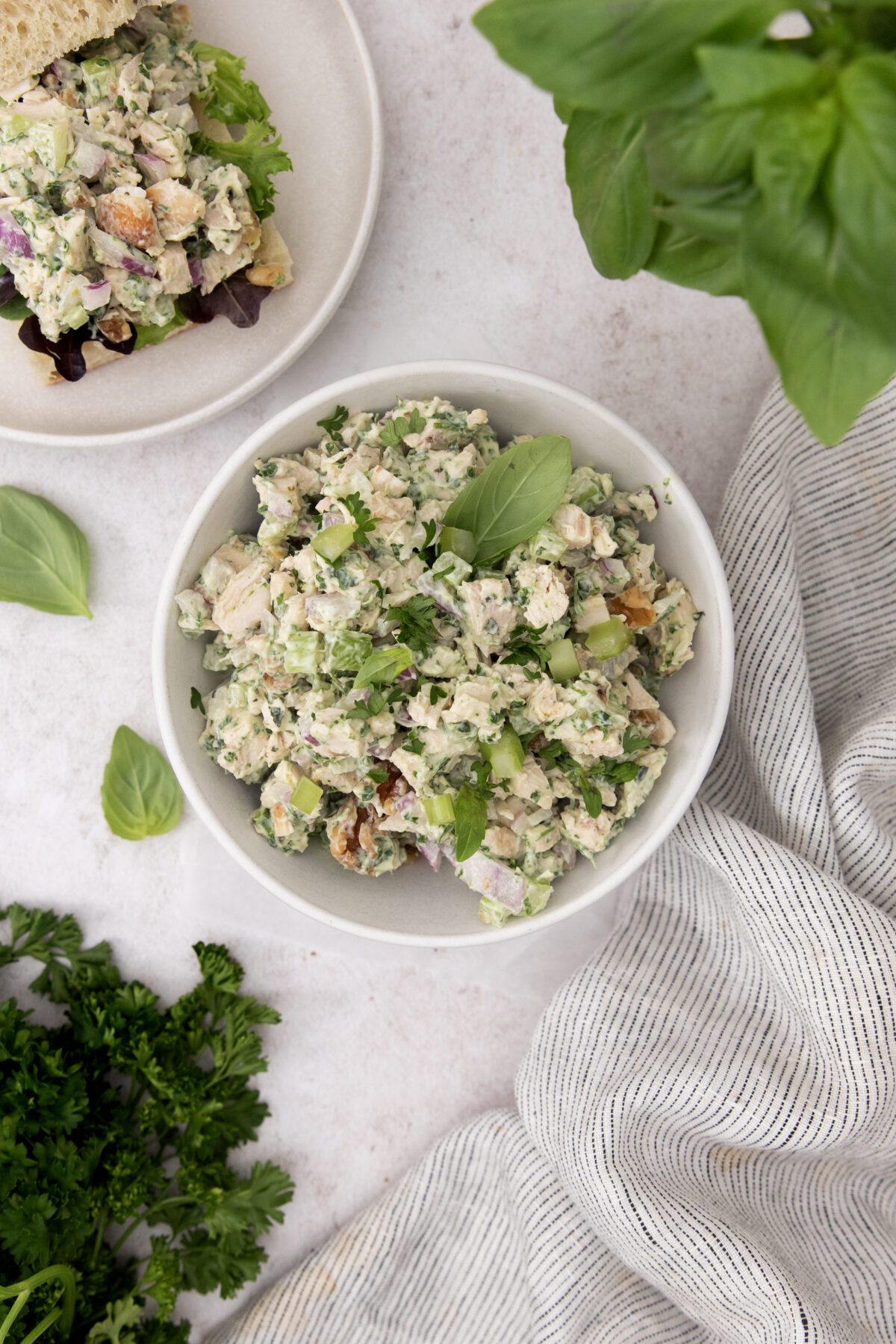 A bowl of creamy chicken salad garnished with fresh herbs sits on a light surface, surrounded by basil leaves, parsley, a striped napkin, and a nearby plate with a sandwich.