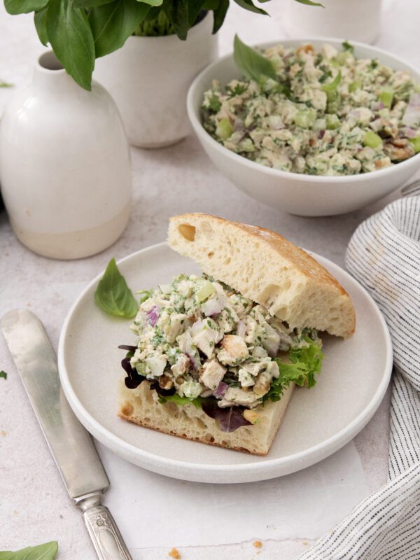 A sandwich on a white plate filled with a chunky salad of chicken, herbs, and vegetables, served on ciabatta bread with lettuce. A bowl of the same salad and a vase of basil are in the background.