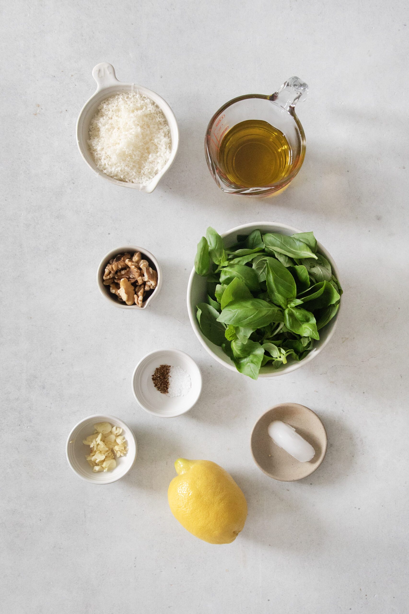 A flat lay of pesto ingredients: a bowl of fresh basil leaves, a small pitcher of olive oil, grated Parmesan cheese, walnuts, lemon, garlic, salt, pepper, and a small ice cube on a light surface.