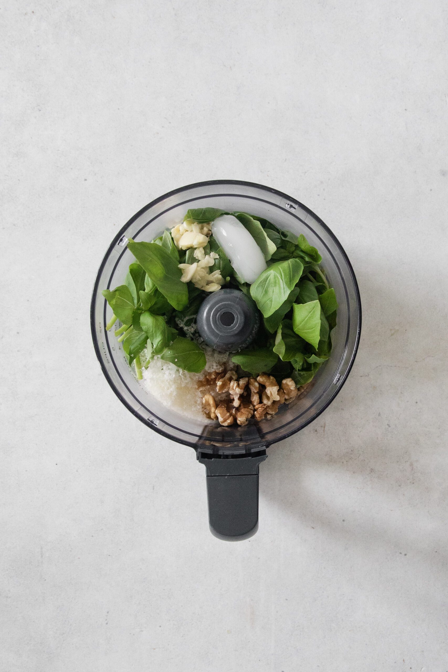 A food processor bowl containing fresh basil leaves, walnuts, grated cheese, garlic cloves, and a chunk of parmesan on a light surface, ready to be blended for pesto.
