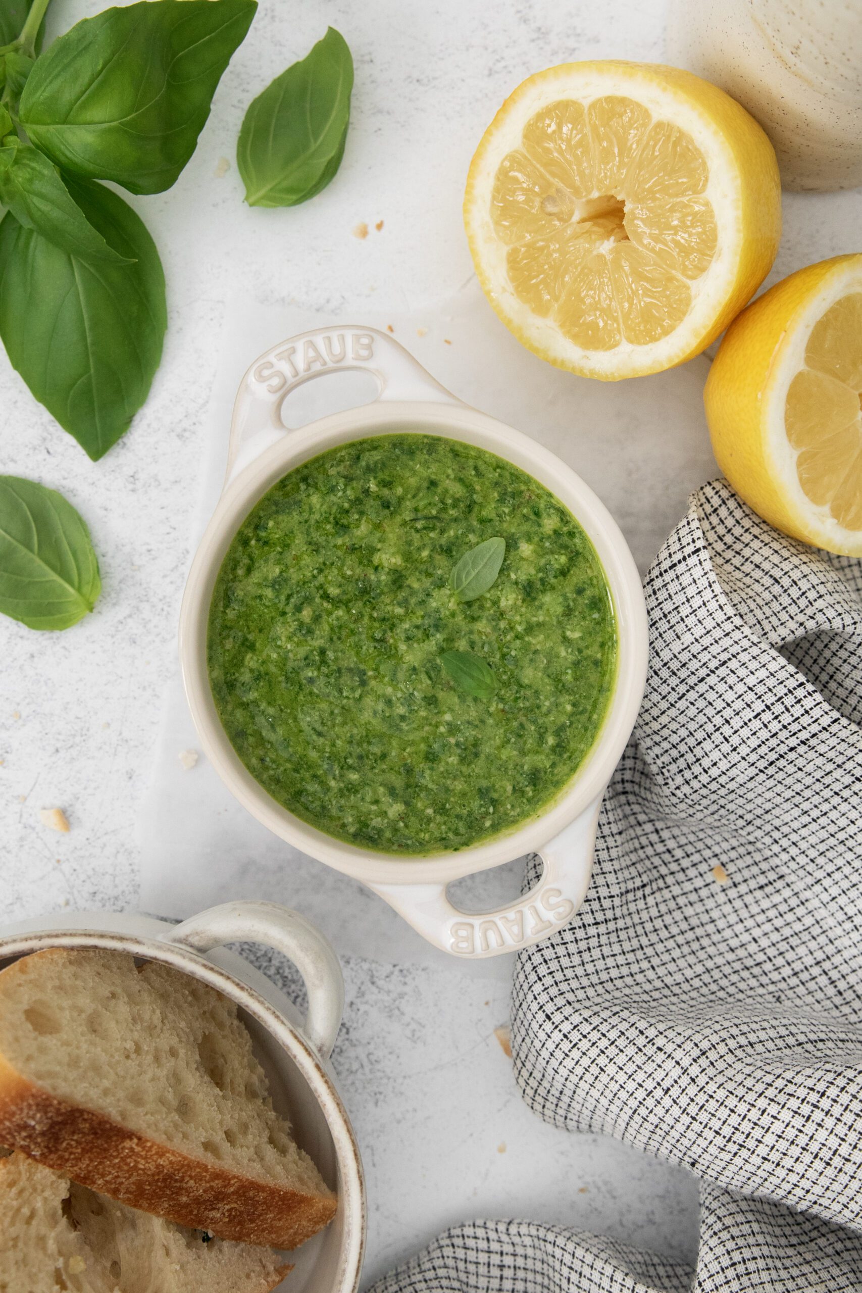 A bowl of green pesto sauce sits on a white surface, surrounded by fresh basil leaves, a halved lemon, a gray checkered cloth, and a dish with slices of bread.