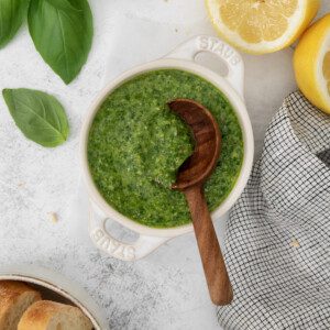 A bowl of green pesto sauce with a wooden spoon, surrounded by fresh basil leaves, a halved lemon, a gray plaid napkin, and slices of bread on a white surface.