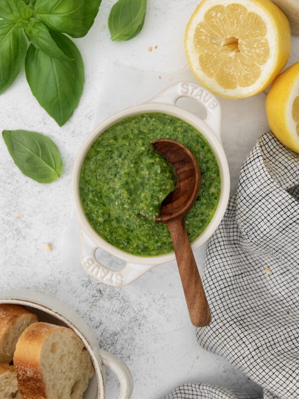 A bowl of green pesto sauce with a wooden spoon, surrounded by fresh basil leaves, a halved lemon, a gray plaid napkin, and slices of bread on a white surface.