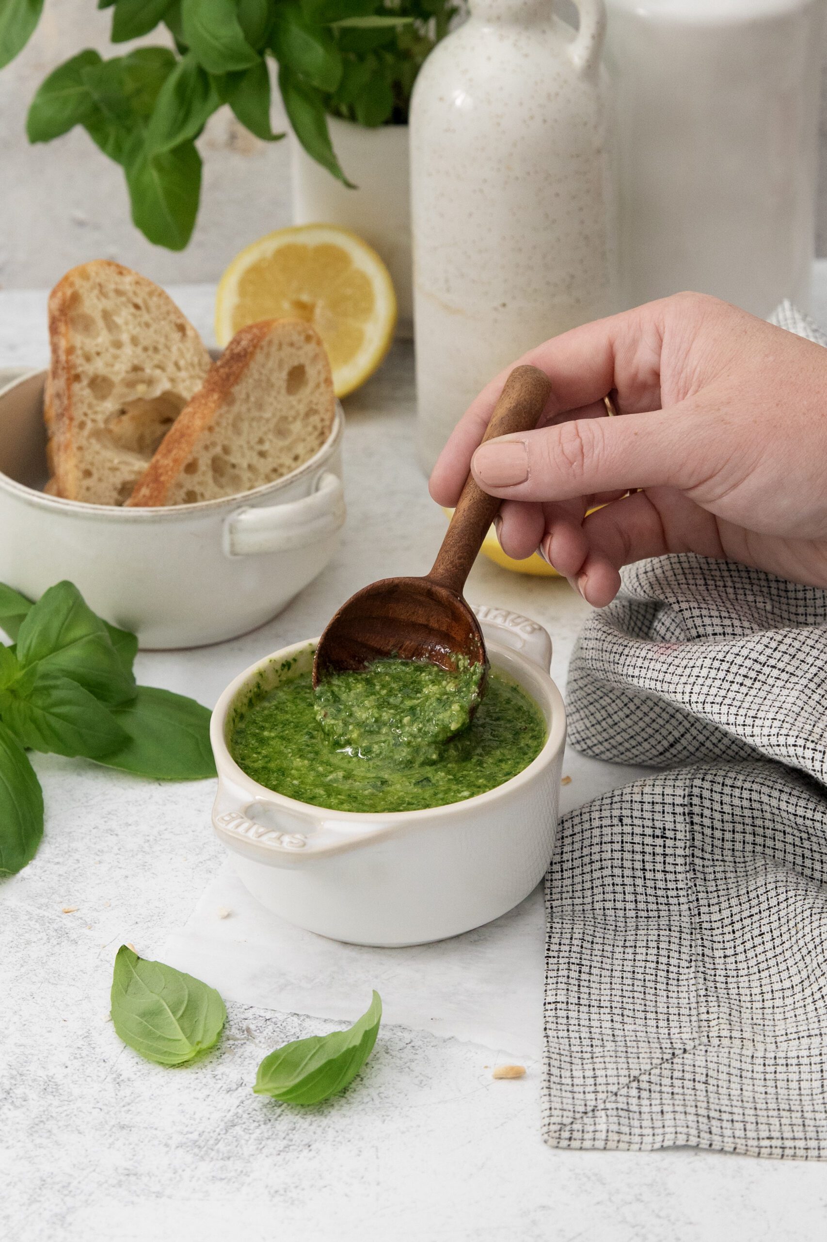 A hand holds a wooden spoon scooping green pesto from a small white bowl. Nearby are fresh basil leaves, a bowl of sliced bread, a lemon half, and a white vase on a light table with a checkered cloth.
