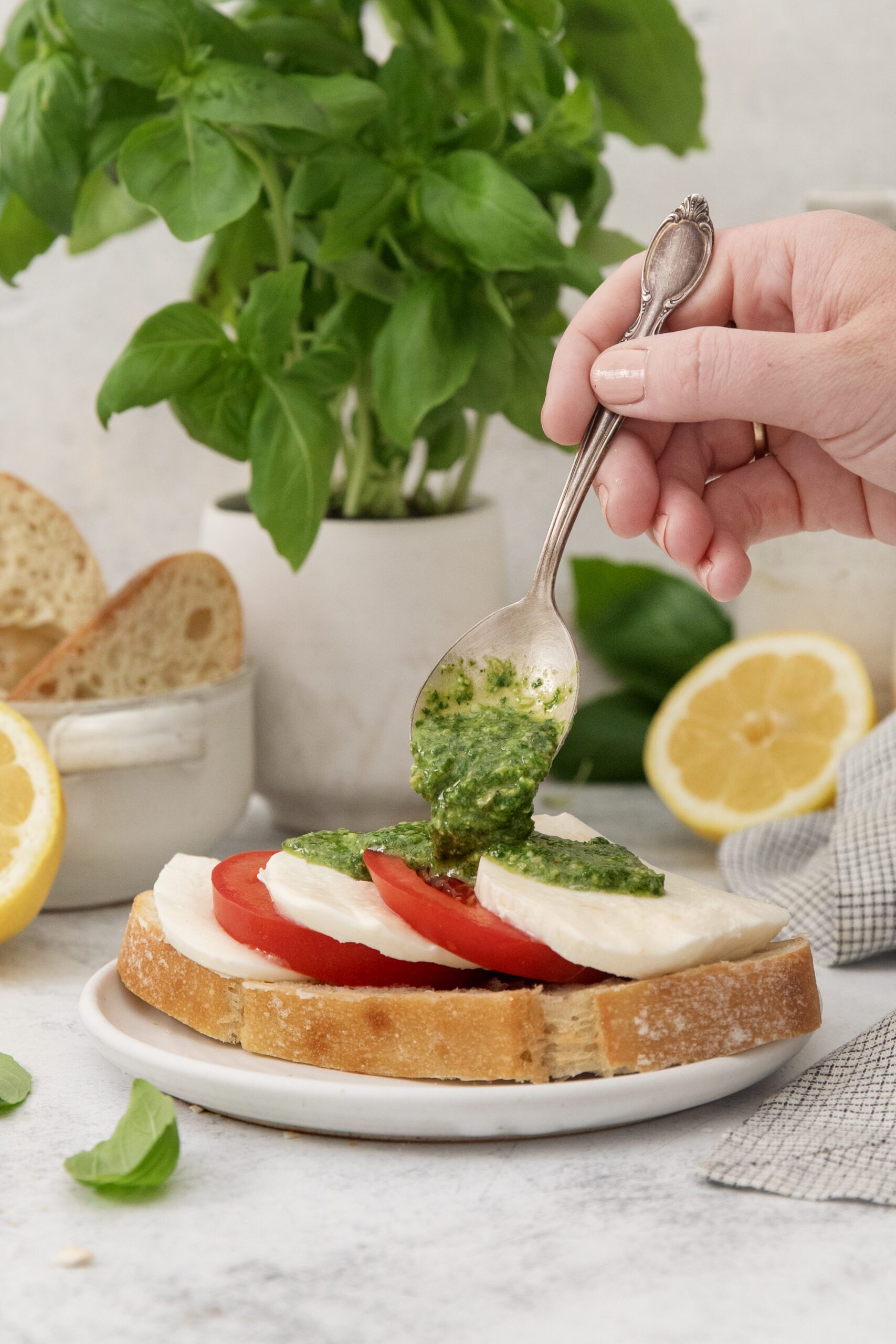 A hand holds a spoon, drizzling green pesto over a slice of bread topped with mozzarella and tomato. Fresh basil, lemon halves, and slices of bread are in the background on a light surface.
