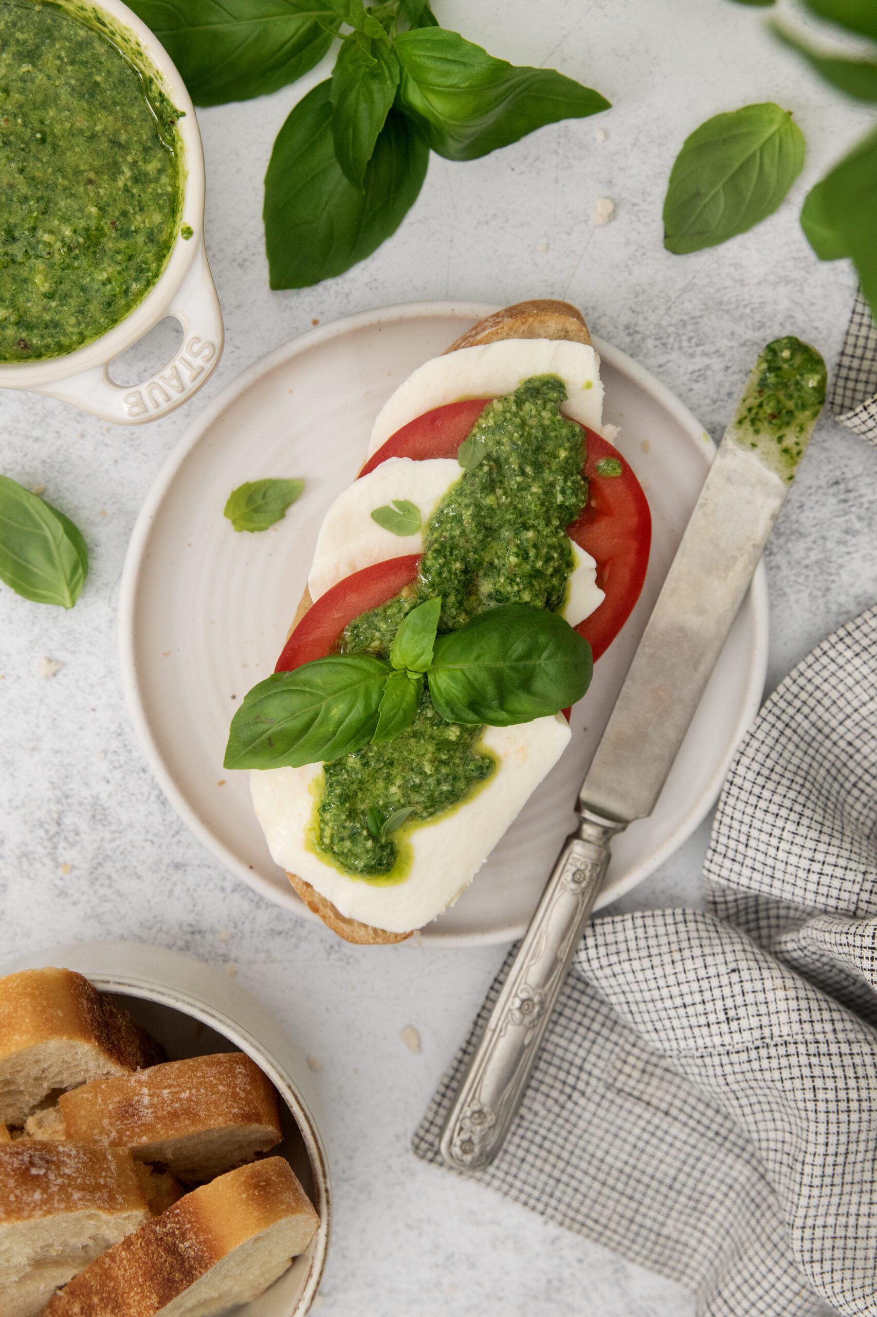 A plate with a slice of bread topped with mozzarella, tomato, pesto, and fresh basil. Nearby are a knife with pesto, a bowl of pesto, basil leaves, a cloth napkin, and sliced bread.
