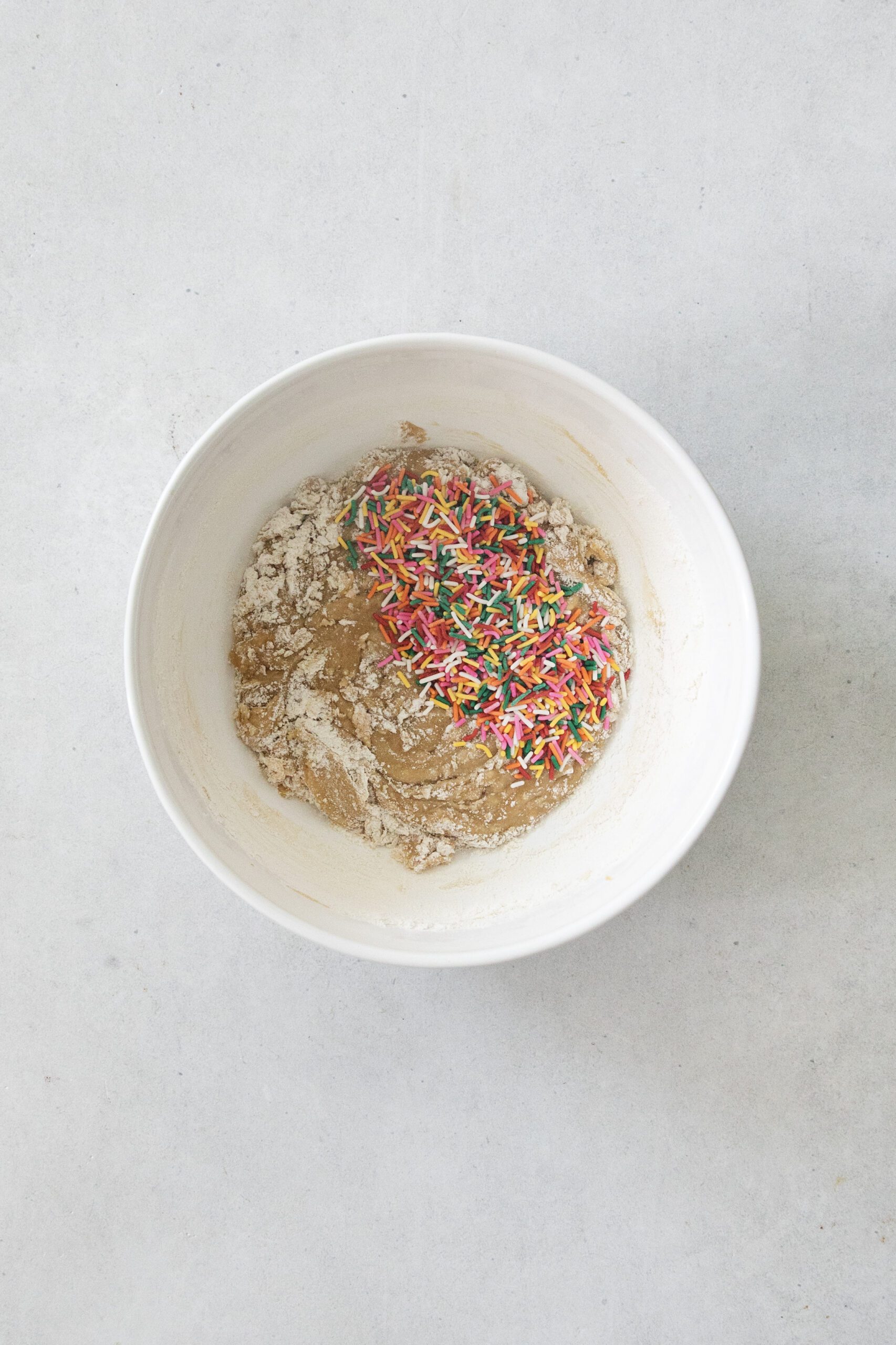 A white mixing bowl containing cookie dough, flour, and a pile of colorful rainbow sprinkles on a light gray surface.