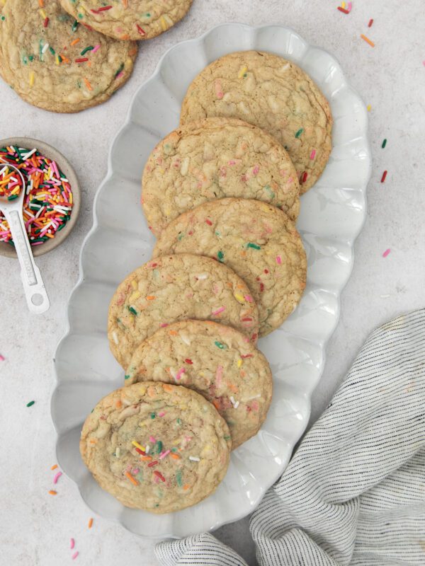 A white scalloped plate holds six large cookies with colorful sprinkles. Nearby, a small dish of sprinkles and a striped cloth are visible on a light surface, with a few scattered sprinkles around.